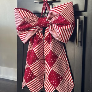 Puede incluir: Un gran lazo decorativo hecho de tela estampada roja y blanca, colgado de un armario oscuro. El lazo presenta un diseño de patchwork con rayas, lunares y cuadros. El lazo está atado con un lazo de tela a juego.