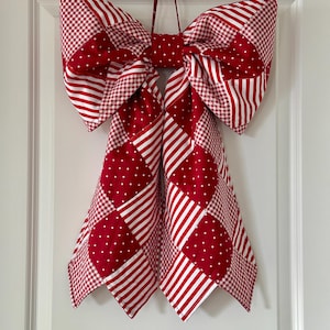 May include: A large fabric bow in red and white patterns hangs on a white door. The bow features a patchwork design with polka dots, stripes, and gingham. The bow is made of fabric and has a loop for hanging.