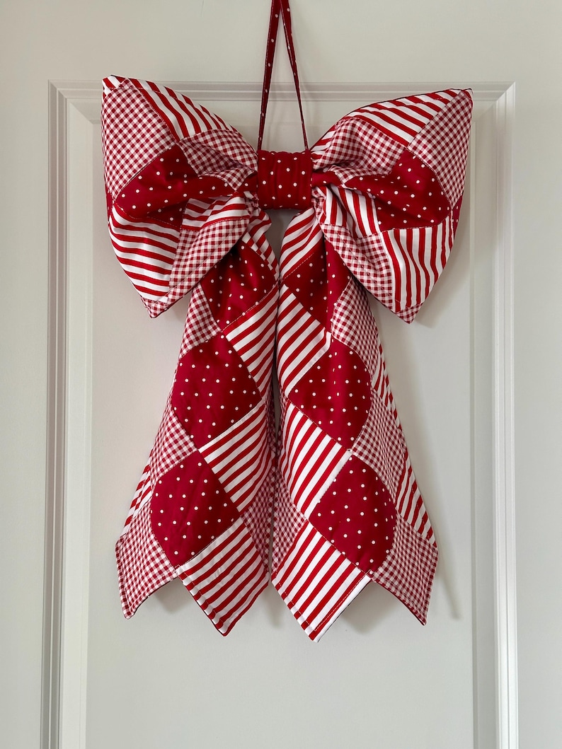 May include: A large fabric bow in red and white patterns hangs on a white door. The bow features a patchwork design with polka dots, stripes, and gingham. The bow is made of fabric and has a loop for hanging.