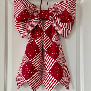 Puede incluir: Un gran lazo decorativo hecho de tela estampada roja y blanca. El lazo presenta patrones de rayas, lunares y cuadros. Est&aacute; dise&ntilde;ado para la decoraci&oacute;n del hogar y est&aacute; colgado de una puerta blanca.