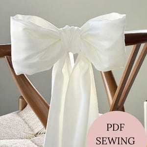 Peut inclure: Un grand nœud en tissu blanc attaché au dossier d'une chaise en bois. Le nœud est fait d'une matière douce et fluide et a une forme pleine et volumineuse. La chaise a une assise tressée. Un cercle rose avec le texte "PDF SEWING PATTERN" est dans le coin inférieur droit.