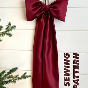May include: A large, burgundy satin bow with long streamers hangs against a white, horizontal-planked wall. The bow is tied at the centre and suspended by a matching ribbon. Text on the right reads "PDF SEWING PATTERN".