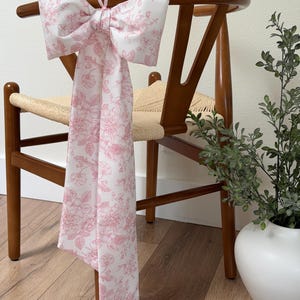 Pode incluir: Um laço decorativo para cadeira feito de tecido branco com um padrão floral rosa. O laço está amarrado ao encosto de uma cadeira de madeira com um assento tecido. O design do laço apresenta uma grande forma de laço com caudas longas.