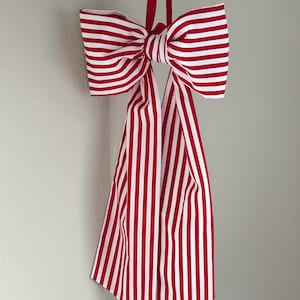 May include: A large decorative bow with red and white stripes, hanging against a neutral gray wall. The bow is made of fabric and has long, striped tails. The bow is suspended by a red ribbon.