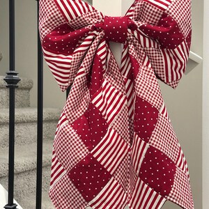Puede incluir: Un gran lazo decorativo hecho de tela estampada roja y blanca, que incluye rayas, lunares y cuadros. El lazo est&aacute; sujeto a un poste blanco, con una escalera en el fondo. Una decoraci&oacute;n festiva.