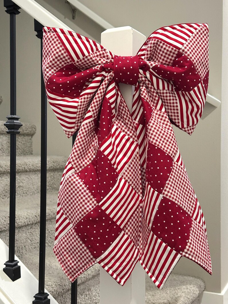 Puede incluir: Un gran lazo decorativo hecho de tela estampada roja y blanca, que incluye rayas, lunares y cuadros. El lazo est&aacute; sujeto a un poste blanco, con una escalera en el fondo. Una decoraci&oacute;n festiva.