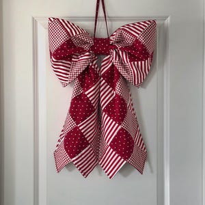 May include: A large, decorative bow made of red and white patterned fabric hangs on a white door. The bow features various patterns, including stripes, polka dots, and gingham. A red fabric loop is used for hanging.