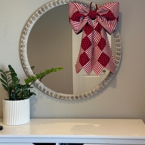 May include: A round mirror with a white, beaded frame features a large red and white patterned bow. A potted green plant sits on a white chest of drawers below the mirror, creating a decorative display.