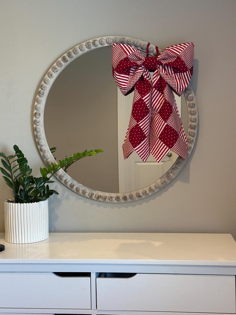 May include: A round mirror with a white, beaded frame features a large red and white patterned bow. A potted green plant sits on a white chest of drawers below the mirror, creating a decorative display.