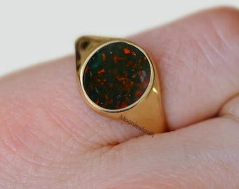 May include: A gold signet ring with a round, dark green stone speckled with red. The ring is on a finger, showcasing the polished gold band and the unique stone setting. The ring is a piece of jewelry.