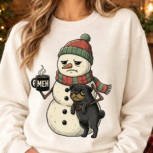 May include: A cream-coloured crewneck sweatshirt featuring a graphic of a grumpy snowman wearing a red and green knit hat and scarf. The snowman holds a black mug with the text MEH and carries a small black pug dog in its stick arms.