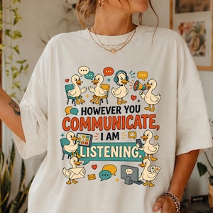 Puede incluir: Una camiseta color hueso con un gráfico de patos de dibujos animados comunicándose de diversas formas. El texto dice "HOWEVER YOU COMMUNICATE, I AM LISTENING". El diseño incluye globos de texto, una computadora, una tableta y un megáfono.
