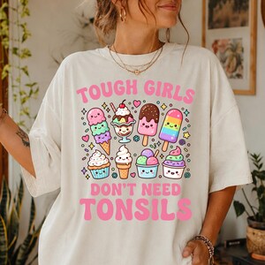 May include: An off-white t-shirt featuring the pink text TOUGH GIRLS DON'T NEED TONSILS and a graphic of various colourful, smiling ice cream treats, including cones, sundaes, and popsicles, surrounded by small stars and hearts.