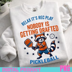 Könnte beinhalten: Weißes Sweatshirt mit einem Waschbären, der Pickleball spielt. Der Waschbär trägt ein orangefarbenes Hemd und blaue Shorts und hält einen Schläger. Der Text lautet "Relax it's rec play, nobody is getting drafted, pickleball". Sterne und ein Pickleball sind ebenfalls abgebildet.