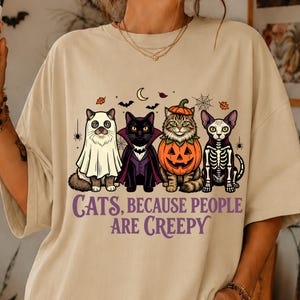 May include: A beige t-shirt featuring a graphic of four cats in Halloween costumes: one as a ghost, one as a vampire, one inside a pumpkin, and one as a skeleton. Below the cats, the text reads: "CATS, BECAUSE PEOPLE ARE CREEPY" in purple.