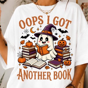 May include: A white t-shirt featuring a graphic of a ghost wearing a purple witch hat, reading an orange book. The ghost is surrounded by stacks of books, jack-o'-lanterns, bats, and spiderwebs. The text reads OOPS I GOT ANOTHER BOOK in orange.