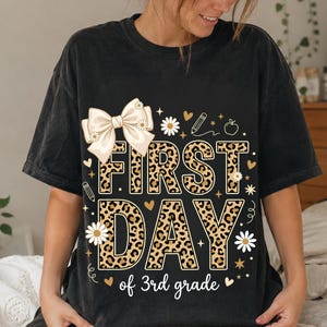 Könnte beinhalten: Ein schwarzes Kurzarm-T-Shirt mit einem Grafikdruck, der den Text "FIRST DAY of 3rd grade" zeigt. Das Design umfasst eine große cremefarbene Schleife, Buchstaben mit Leopardenmuster, weiße Gänseblümchen, einen Bleistift und ein Apfelsymbol.