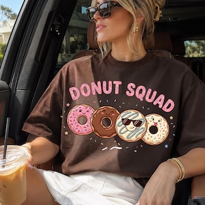 以下が含まれることがあります： ダークブラウンの半袖Tシャツ。ピンク色の文字でDONUT SQUADと書かれ、その下に4つのドーナツのイラストが描かれています。ドーナツは、スプリンクル付きのピンク色、チョコレート色、青いアイシングとサングラス付き、そして白いドーナツの4種類です。