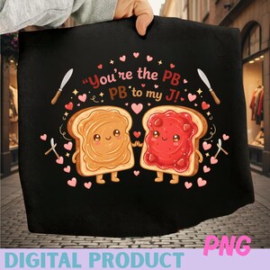 以下が含まれることがあります： ピーナッツバターとゼリーを塗った2枚の漫画のパンのスライスが描かれた黒いグラフィック。テキスト「You're the PB to my J！」は、ハートとナイフで囲まれています。