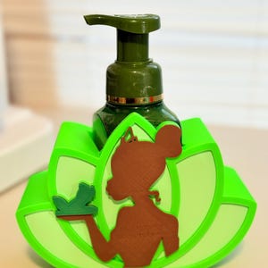May include: A green soap dispenser with a brown silhouette of a woman holding a frog, set in a bright green lotus flower-shaped holder. The dispenser has a dark green pump and a gold band.
