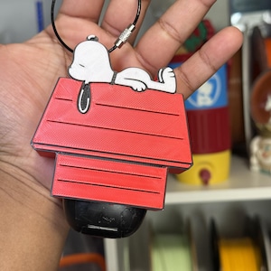 May include: A 3D-printed keychain featuring a white Snoopy figure sleeping on top of a red doghouse. The design includes a black metal wire loop for attachment, and the entire piece is mounted onto a black protective case for wireless earbuds.