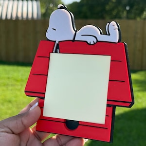 Puede incluir: Un soporte para notas de plástico rojo y negro con forma de caseta de perro, con un perro de dibujos animados blanco tumbado en el tejado. El centro de la estructura roja contiene un bloc de notas adhesivas cuadrado de color amarillo pálido.