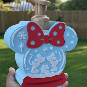 May include: A light blue soap dispenser shaped like a mouse head with a red bow and a red base. The front features white silhouettes of two mice facing each other with a gift box, surrounded by white snowflake and star accents and a tan pump.