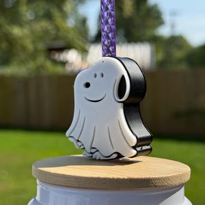 May include: A white and black ghost-shaped straw topper featuring a smiling face, attached to a purple translucent twisted straw. The topper sits on a light-colored wooden lid that rests on top of a white cylindrical container.