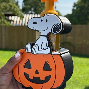 May include: A soap dispenser holder featuring a white Snoopy character sitting atop a bright orange jack-o'-lantern pumpkin. The holder is constructed from black material and includes a yellow pump mechanism extending from the top of the container.