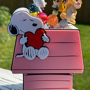 May include: A pink doghouse-shaped pen holder featuring a Snoopy character holding a red heart on the front. Several decorative pens with character toppers, including Alice in Wonderland and a rabbit, are placed inside the container.