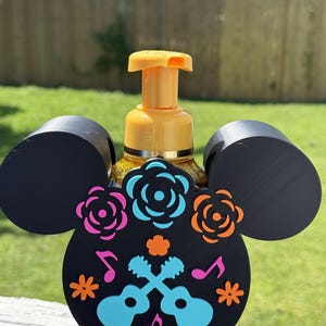 Pode incluir: Um suporte de plástico preto para dispensador de sabonete em forma de cabeça de rato, com detalhes decorativos coloridos, incluindo flores azul-claras e laranja, duas guitarras cruzadas azul-claras e notas musicais cor-de-rosa, segurando um frasco de sabonete amarelo.