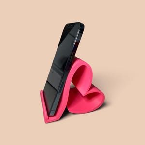 May include: A hot pink phone stand with a curved, heart-like design. A black smartphone is placed in the stand, angled for viewing. The stand is made of a smooth material and sits on a neutral background.