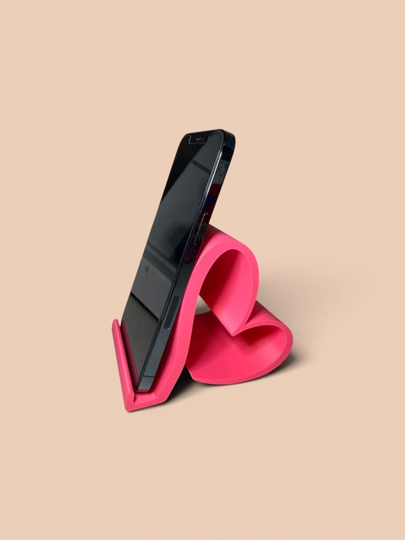 May include: A hot pink phone stand with a curved, heart-like design. A black smartphone is placed in the stand, angled for viewing. The stand is made of a smooth material and sits on a neutral background.