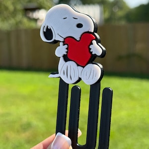 May include: A black, white, and red 3D printed bookmark featuring a cartoon dog character holding a red heart. The character is attached to a large, black, U-shaped paperclip design. The object has a textured surface finish throughout.