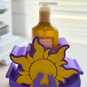 May include: A purple and yellow decorative soap dispenser holder. The holder features a yellow sun design with a silhouette of two figures. A bottle of soap sits inside the holder.