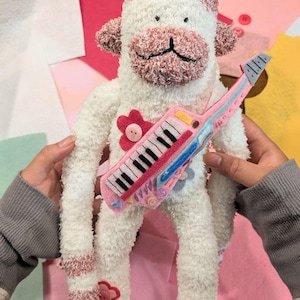 以下が含まれることがあります： 白とダスティピンクのふわふわした生地で作られた靴下猿のぬいぐるみ。頭と脚にボタンが中央についたピンクのフェルトの花が飾られています。黒と白の鍵盤がついたピンクと青のフェルト製のキーターを持っています。