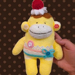 May include: A yellow sock monkey plush toy featuring a red pom-pom and white felt topping on its head. It has a clear button on one ear and a red-and-white checkered button on the other. A pink button, white lace trim, and a blue bow decorate its torso.