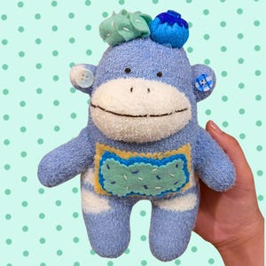 May include: A plush sock monkey toy made of fuzzy light blue fabric with a white face, featuring a rectangular felt patch on the chest, a blue berry and green leaf accent on the head, and two mismatched buttons for ears in white and blue plaid.
