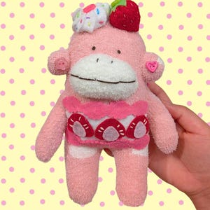 May include: A pink plush sock monkey toy featuring a white face, a red strawberry and a white frosted cupcake on its head, and a pink jumper with a strawberry pattern. The monkey has a small pink button on each ear, one round and one heart-shaped.