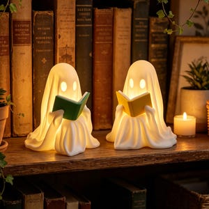 May include: Two white ghost-shaped lamps made of smooth material, each featuring glowing eyes and holding a small book. One ghost holds a green book, while the other holds a yellow book. The lamps emit a warm, soft light from within their forms.