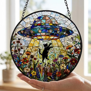 Pode incluir: Um apanhador de sol circular em vitral com um gato preto a alcançar um OVNI azul e metálico que paira sobre um jardim vibrante cheio de flores multicoloridas, tudo dentro de uma moldura de metal preta fina com uma corrente de suspensão.