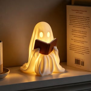 May include: A glowing, off-white ghost-shaped lamp made of smooth material, featuring two oval eye cutouts and holding a small, dark brown open book. The lamp emits a warm, soft light from within, illuminating its draped, fabric-like form.