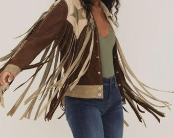 Könnte beinhalten: Braune Wildlederjacke mit beigen Fransen und Sternen. Die Jacke hat einen beigen Kragen und Manschetten, kombiniert mit einem grünen Oberteil und blauen Jeans. Die Jacke ist im Western-Stil gehalten.
