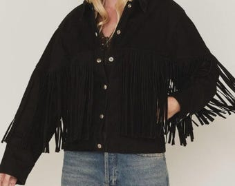 May include: Black denim jacket with long fringe detailing across the chest and sleeves. The jacket has a button-up front and a classic collar. The model is wearing blue jeans. A stylish, western-inspired outerwear piece.