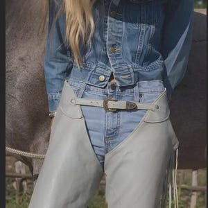 May include: A light blue denim jacket and jeans outfit. The ensemble features a light grey leather belt with a decorative buckle and matching chaps with fringe detailing. The model is wearing a cowboy hat.