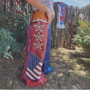 Op de afbeelding: Rode, witte en blauwe leren chaps met een patriottisch ontwerp. De chaps hebben de Amerikaanse vlag, drie omrande sterren en rode franjes. Het bovenste gedeelte is bruin met een decoratief golfpatroon.