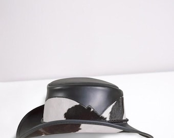 Puede incluir: Sombrero de vaquero de piel de vaca en blanco y negro con corona y ala de cuero negro. El sombrero presenta un ribete trenzado decorativo alrededor de la base de la corona. Diseño de estilo western clásico.