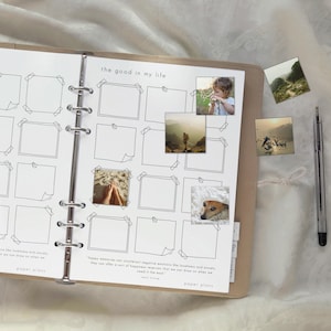 Könnte beinhalten: Ein Ringbuchplaner mit beigem Einband und einer Seite mit der Überschrift the good in my life, leeren quadratischen Rahmen und vier kleinen quadratischen Fotos. Ein silberner Stift liegt rechts neben dem Planer auf weißem Stoff.