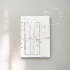 May include: A white paper planner with a black outline of a smartphone, designed as a screentime tracker. The planner is tied with a string and includes the text "screentime tracker" and "paper plans".