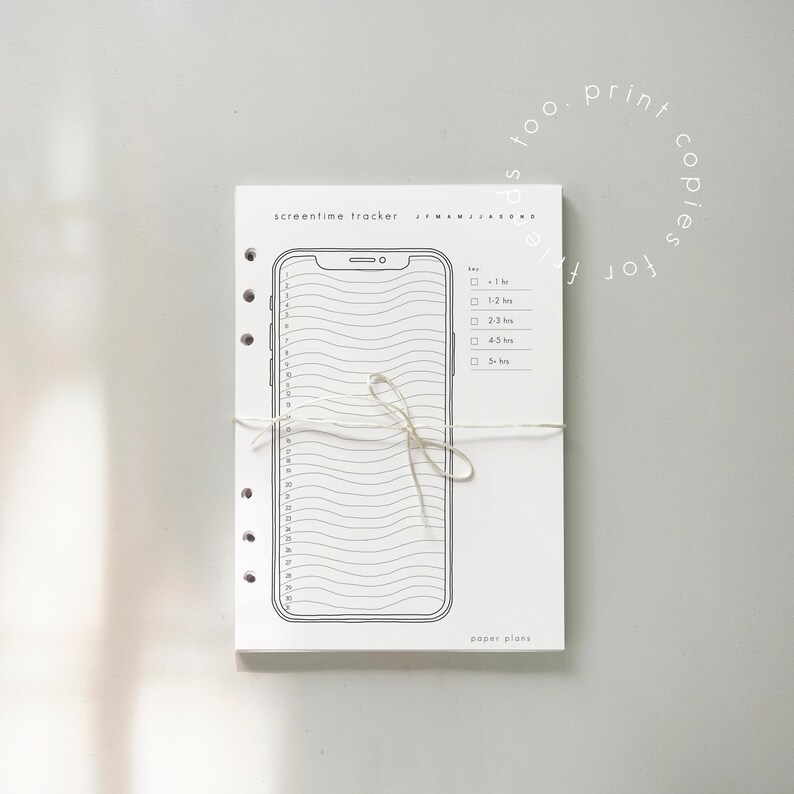 May include: A white paper planner with a black outline of a smartphone, designed as a screentime tracker. The planner is tied with a string and includes the text "screentime tracker" and "paper plans".